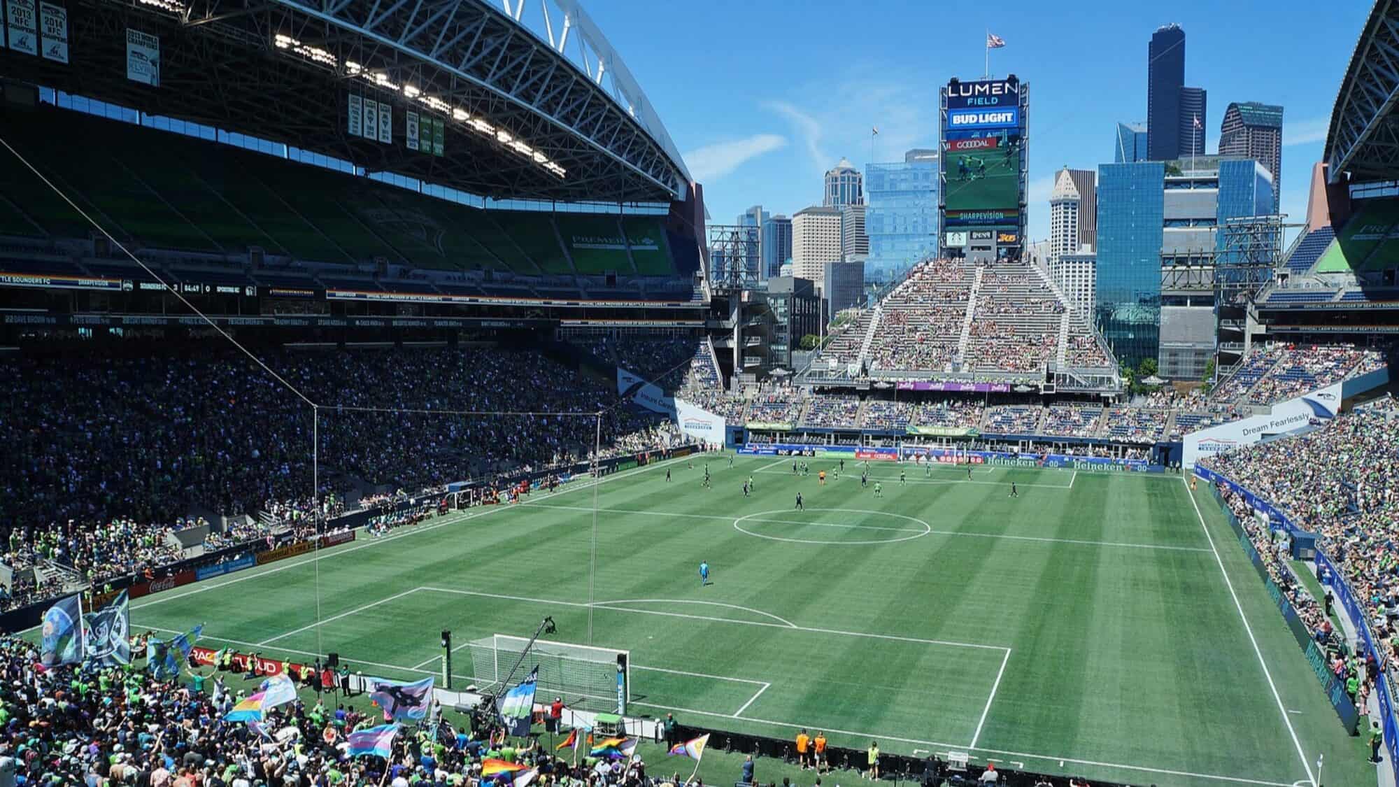 A large soccer stadium filled with fans and World Cup excitement, featuring a green field and a city skyline in the background under a clear blue sky.