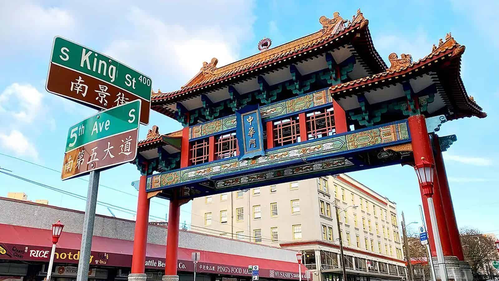 A traditional Chinese archway stands at the intersection of S King St and 5th Ave S, marking one of Seattle’s cultural landmarks, with street signs visible in front and buildings in the background.
