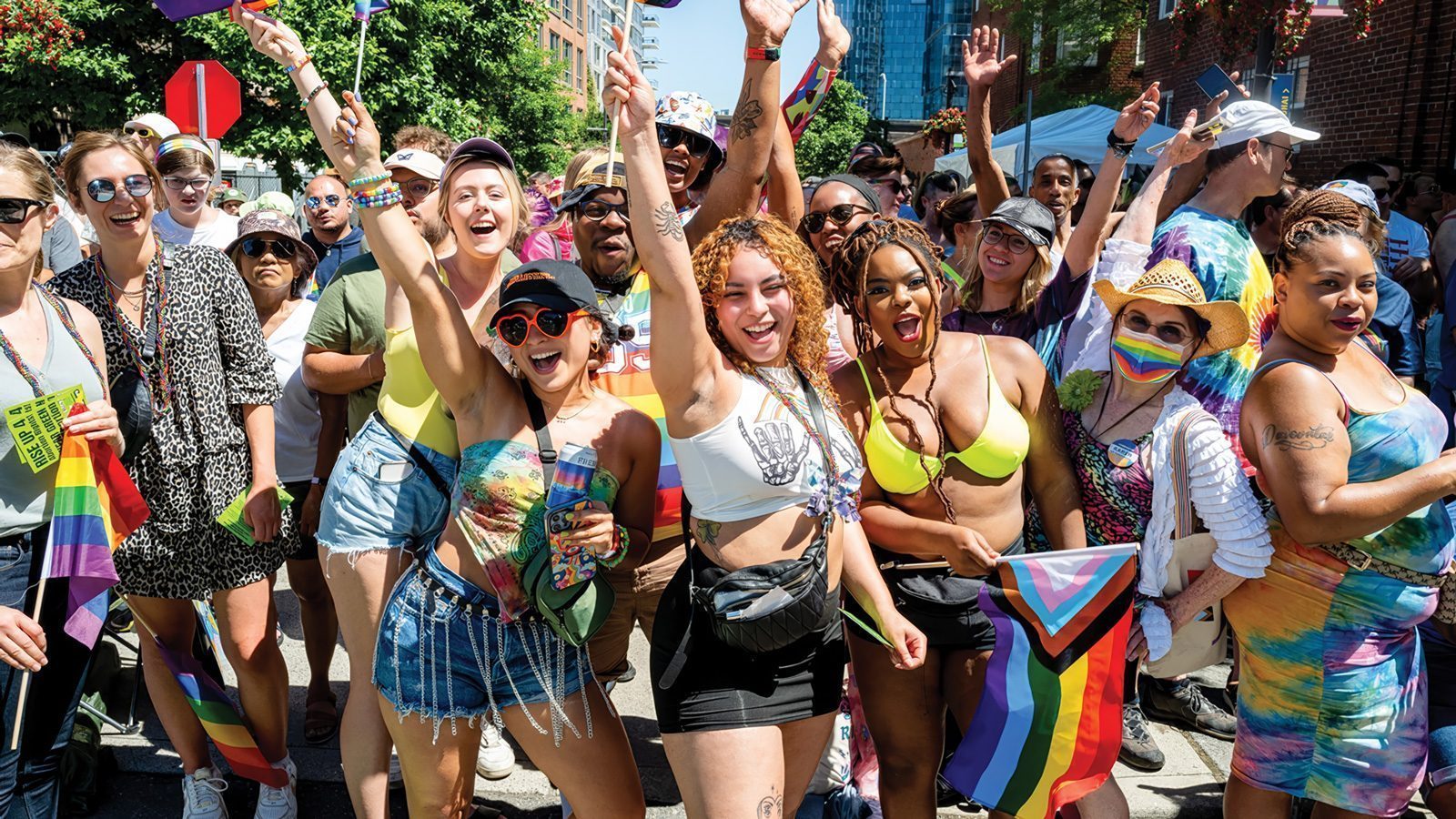 A lively group of people at Seattle Pride, wearing colorful attire and holding rainbow flags, revamps the celebration with smiles and raised arms.