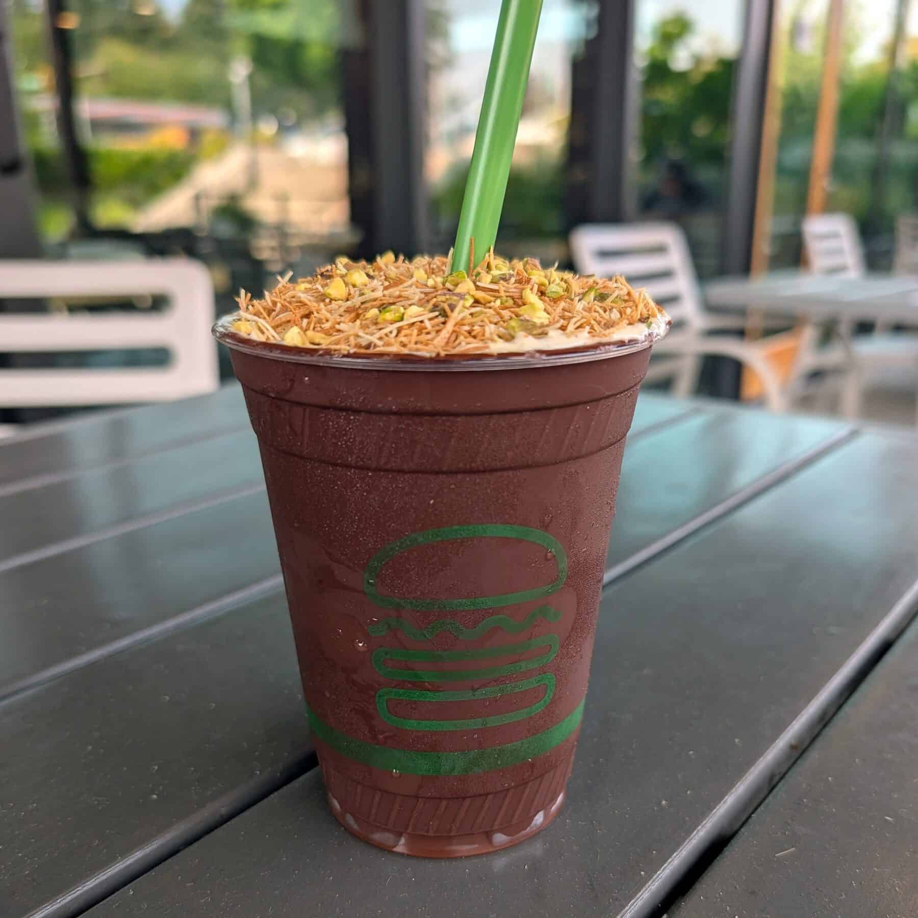 A chocolate milkshake topped with chopped nuts and shredded pastry, served in a plastic cup with a green straw, perfect for enjoying seasonal eating and savoring what to eat in September on an outdoor table.