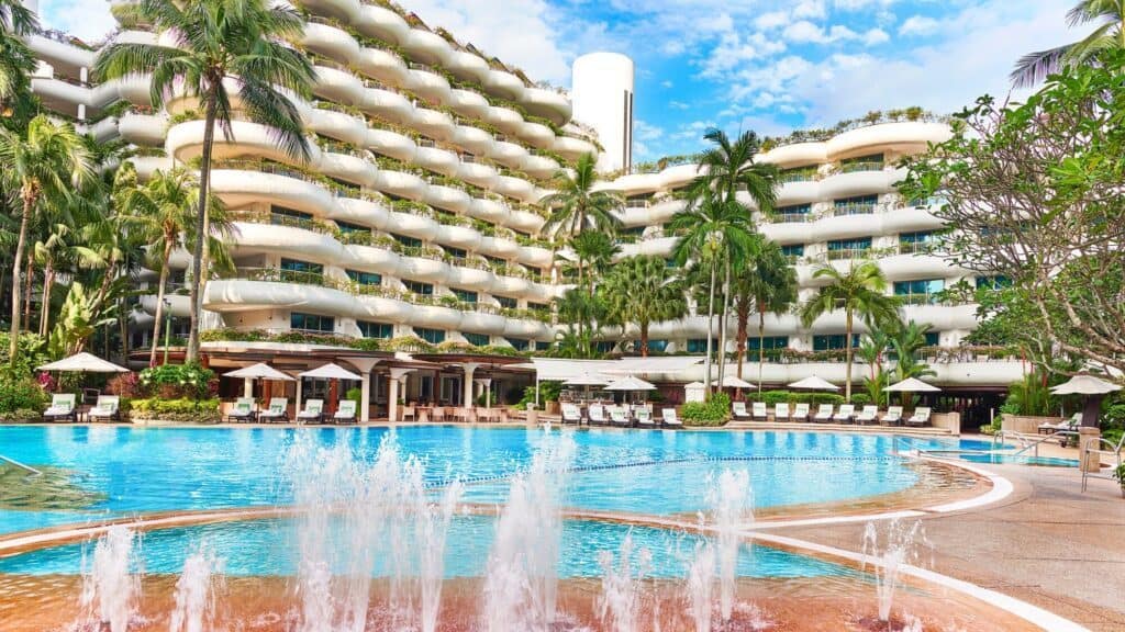 A large outdoor swimming pool with fountains, surrounded by palm trees, sun loungers, and a modern multi-story hotel building in the background.