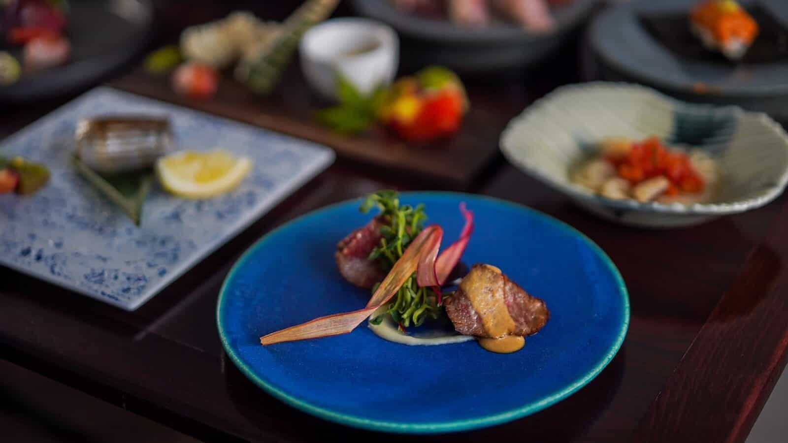 A blue plate with seared meat, sauce, and garnish is in focus; other assorted dishes are blurred in the background on a dark table.