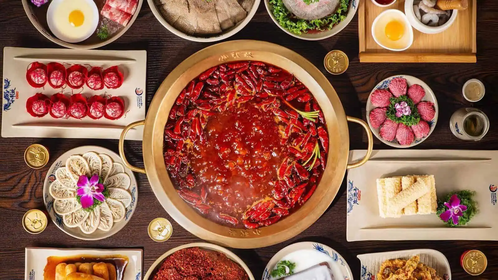 A large pot of spicy hotpot with vegetables, chili peppers, and local tofu is surrounded by various raw meats, seafood, and dipping sauces on a table.