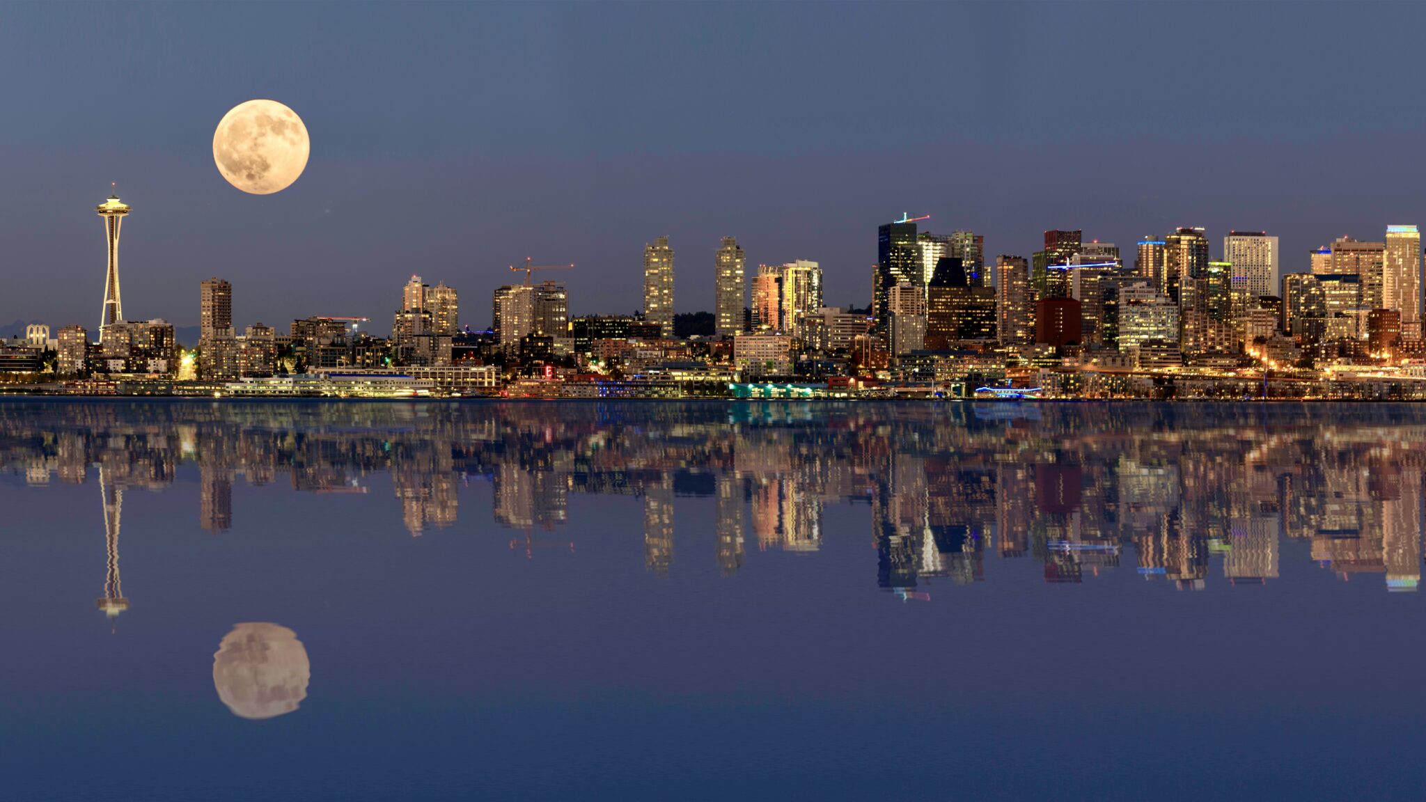 Seattle city skyline at dusk with the Space Needle, city lights, an October Moon, and reflections on calm water.