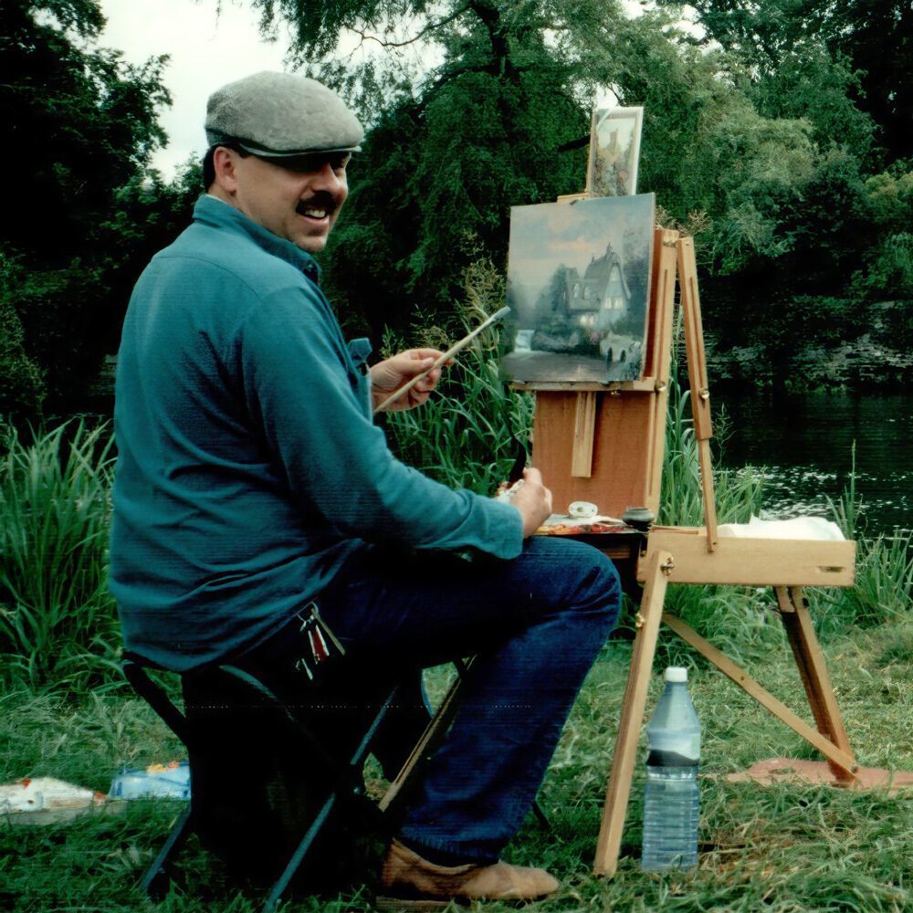 A man sits on a stool outdoors painting a landscape scene on canvas, with his art supplies and a water bottle beside him. Trees and grass form the background—it's a Must List moment for 07-03-2025.