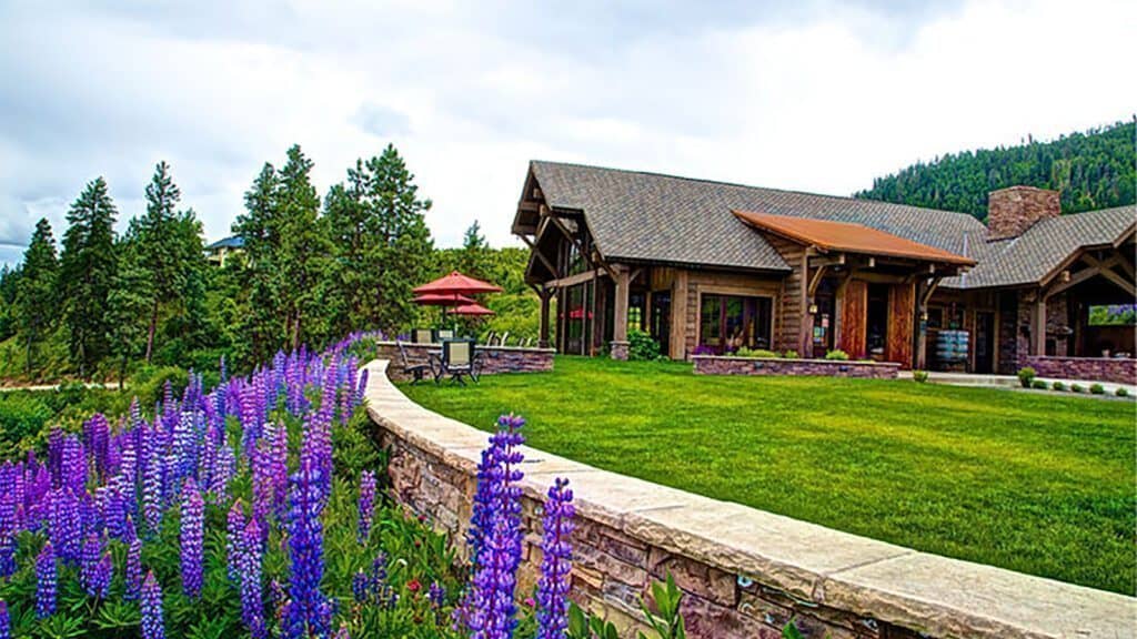 Modern house with a stone facade and wooden roof, surrounded by a well-manicured lawn and purple flowers, set against a backdrop of trees and hills in Hidden Washington.