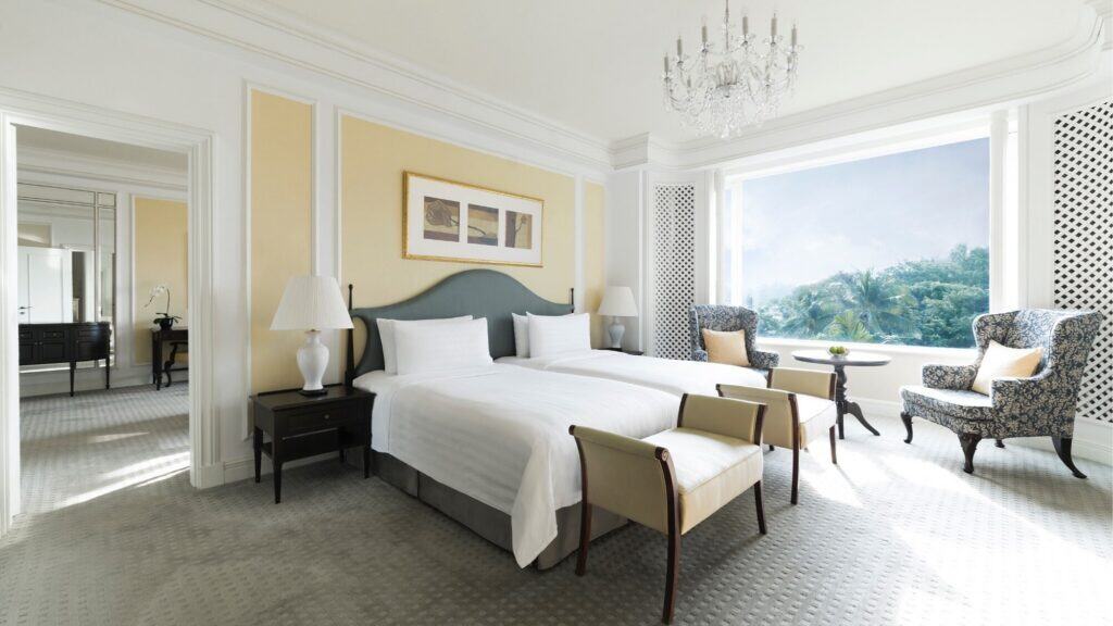 A bright, spacious hotel room with two beds, armchairs by a large window, a chandelier, neutral decor, and a garden view.