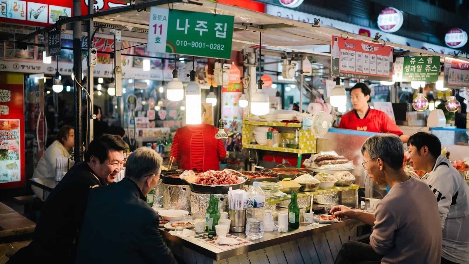 People are seated at a bustling street food stall in an indoor market, enjoying various dishes and drinks under bright lights, with the stall displaying signs in Korean—a taste of one of the Ten Top Travel Destinations from Seattle for 2025.