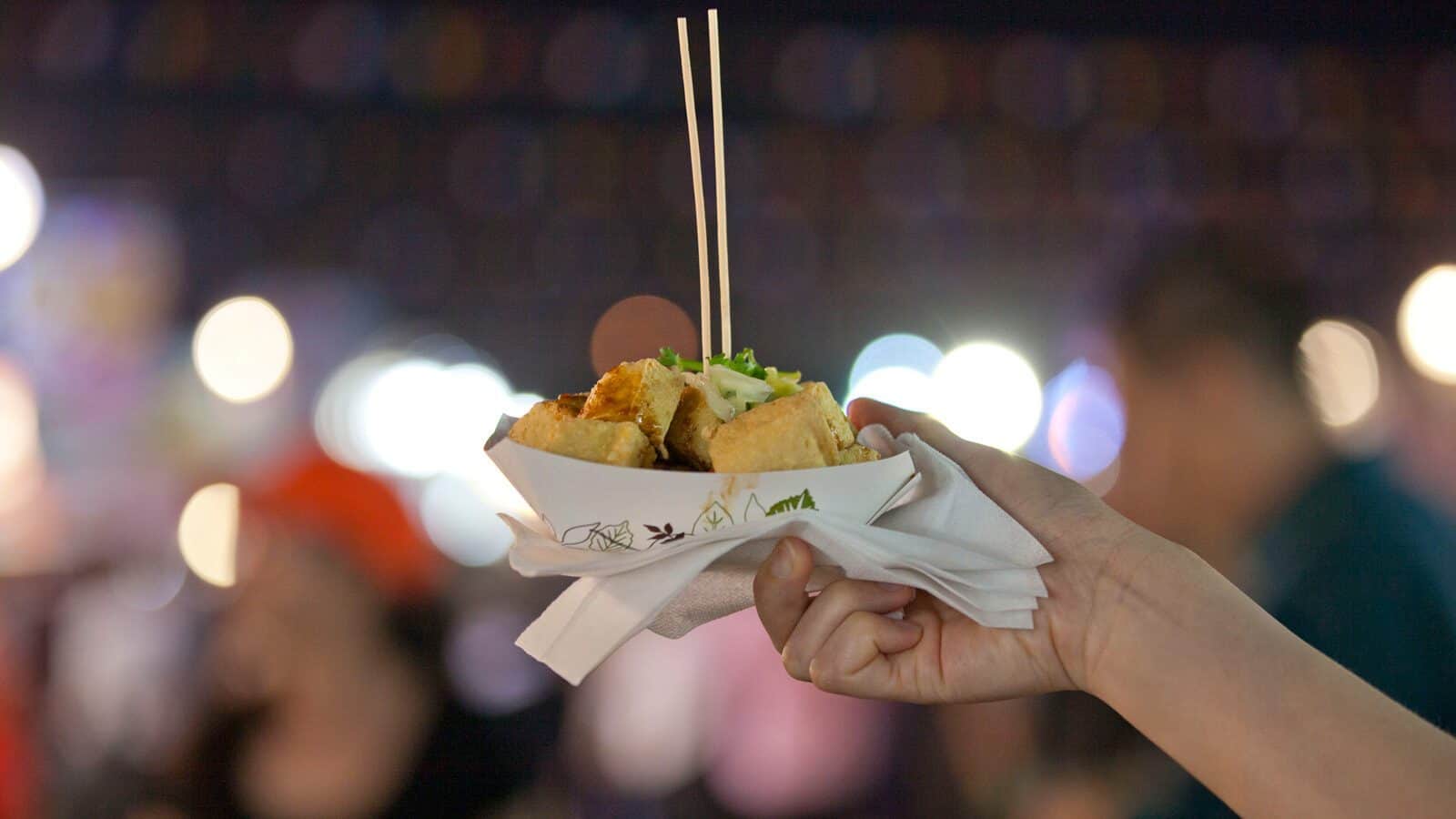 A hand holds a paper tray of Taiwanese Street Food with chopsticks, set against a blurred background of people and lights at night—capturing the vibrant flavors and ambiance of Stateside markets.