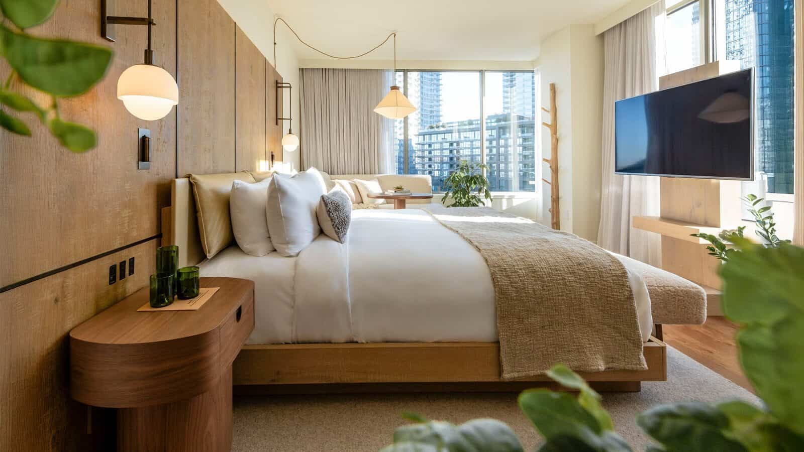 Modern hotel room at 1 Hotel with a large bed, wooden furnishings, neutral decor, plants, wall-mounted TV, and floor-to-ceiling windows overlooking South Lake Union city buildings—a true hidden gem for urban explorers.
