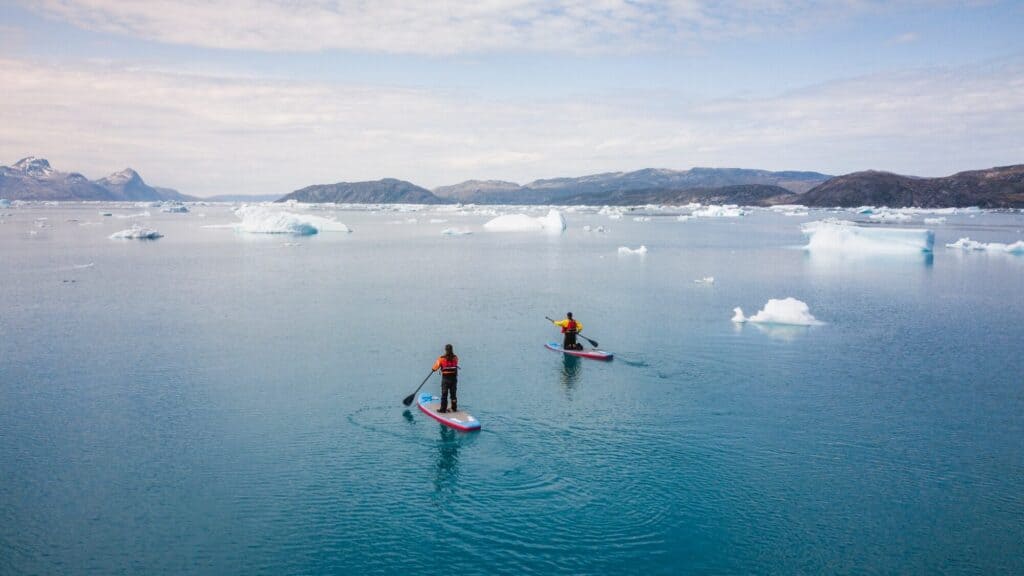 Two people paddleboarding on calm, icy blue water surrounded by floating icebergs with mountains visible in the distance under a partly cloudy sky.