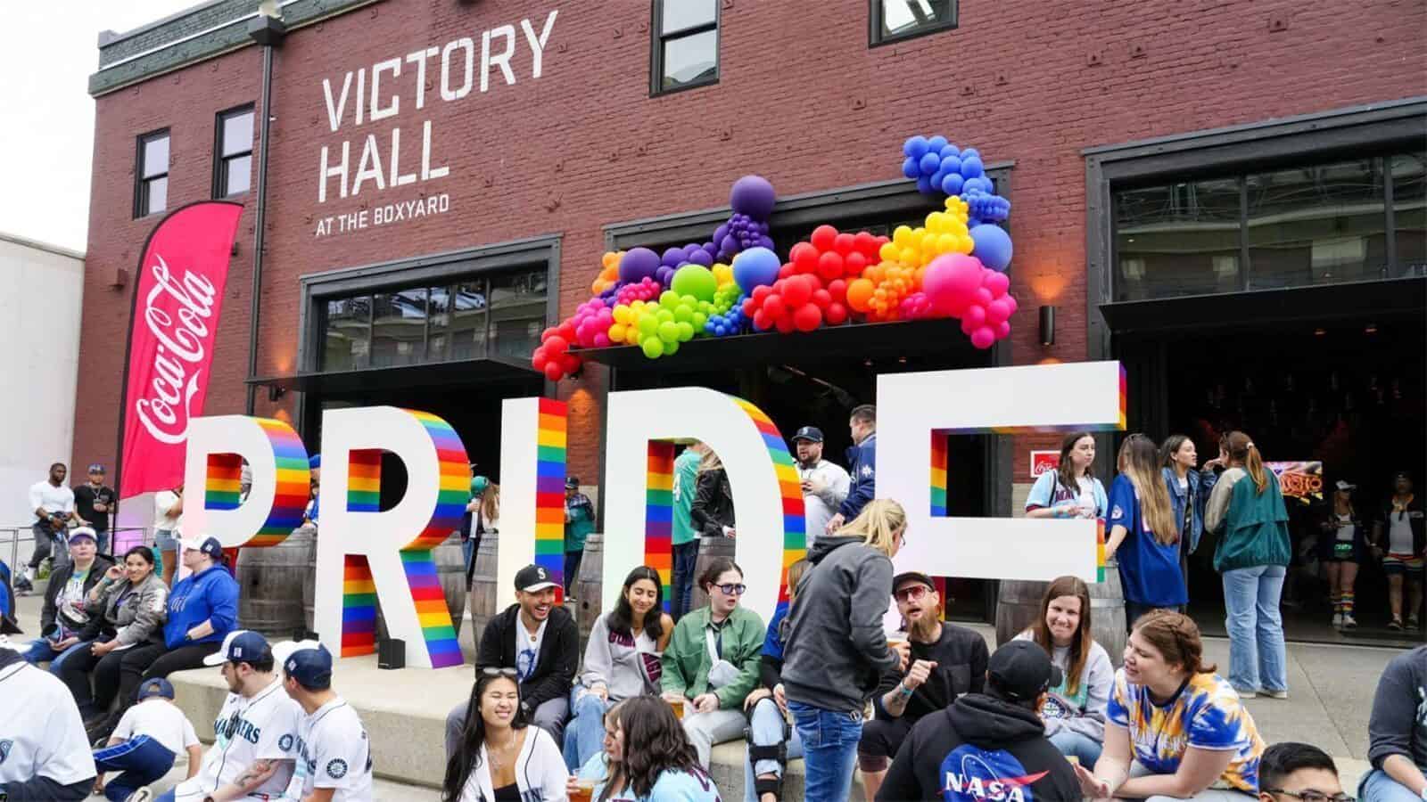 People gather in front of large rainbow-colored "PRIDE" letters and balloon decorations outside Victory Hall, where vibrant celebrations honor Seattle’s rich LGBTQIA+ history at this lively public event.