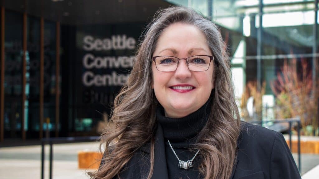 A woman with long brown and gray hair, glasses, and a black turtleneck smiles in front of the Seattle Convention Center building.