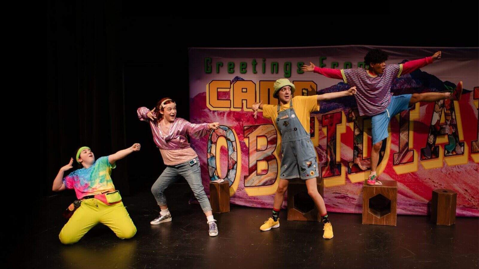 Four people in colorful, playful outfits pose energetically on stage before a backdrop reading “Greetings from Camp,” evoking vibrant celebrations that honor Seattle’s rich LGBTQIA+ history with a lively, camp-themed performance.