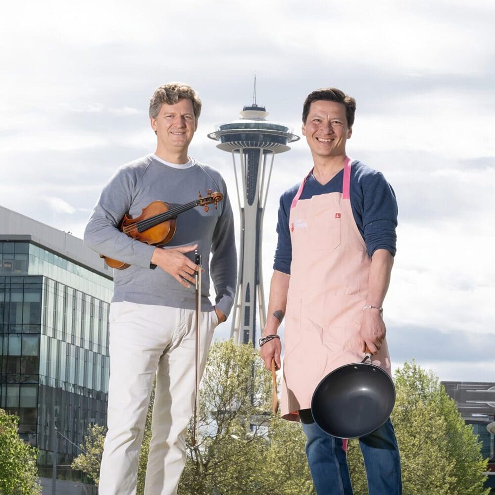 Two men stand outdoors in front of the Space Needle; one holds a violin and bow, the other wears an apron and holds a frying pan and rolling pin. Don’t miss this Must List moment—save the date: 07-24-2025!.