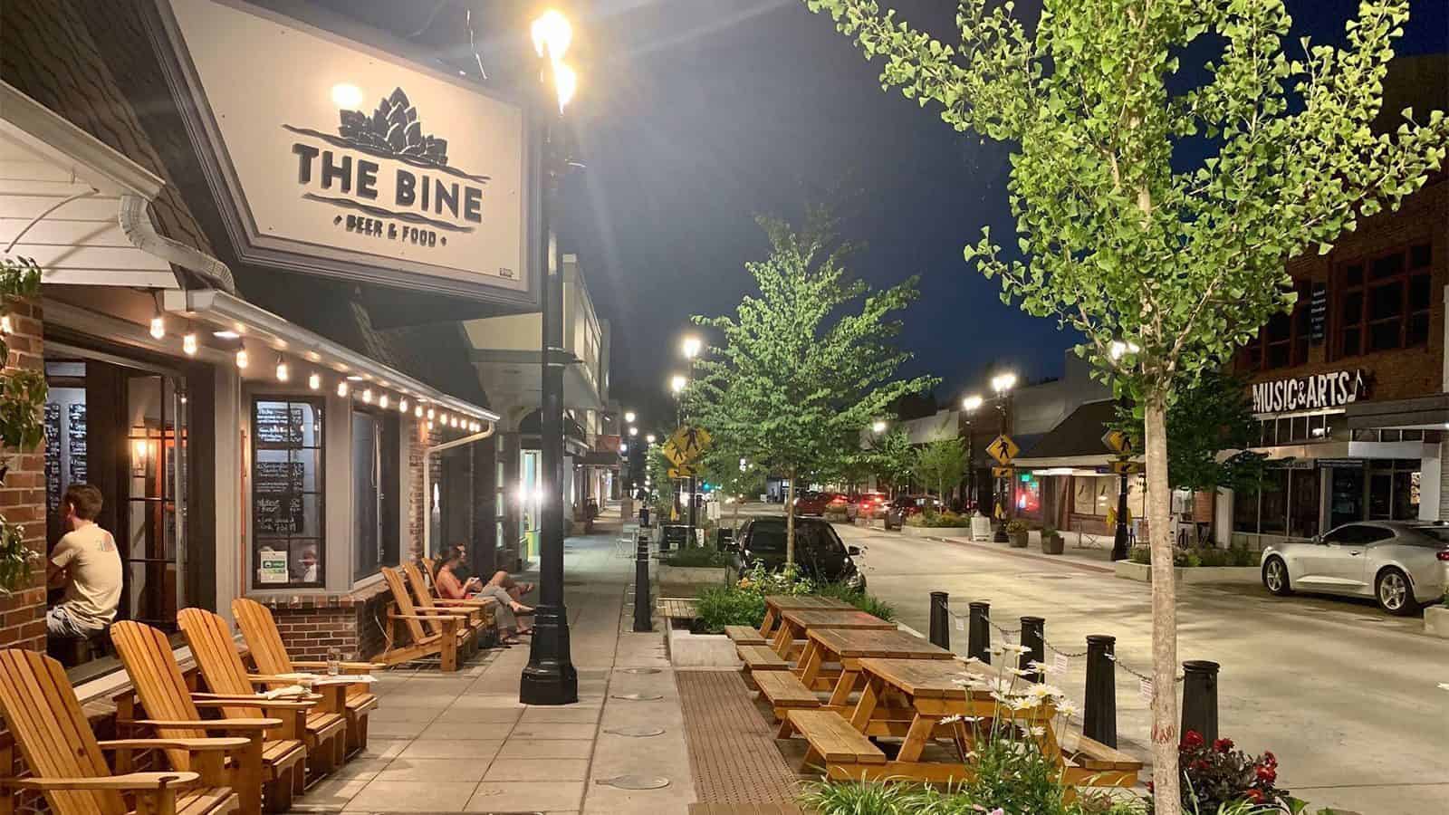 Nighttime city street scene in Bothell with restaurant signage for "The Bine Beer & Food," wooden benches outside, streetlights, and pedestrians. Trees line the sidewalk, and cars are parked along the street. Welcome to Bothell, one of Hidden Washington's gems.