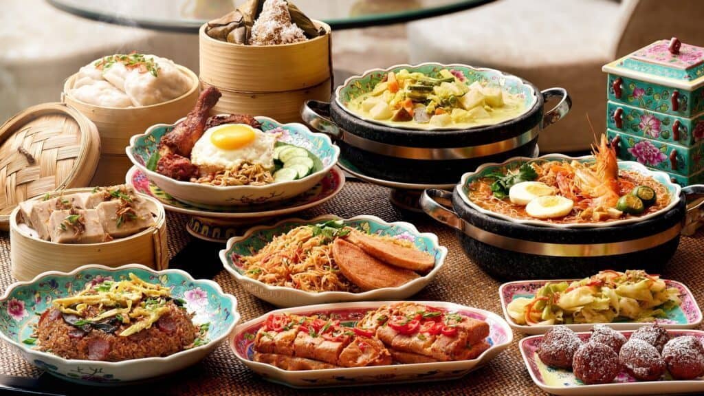 A variety of colorful Asian dishes including noodles, steamed buns, rice, vegetables, and desserts, served in ornate bowls and plates on a table.