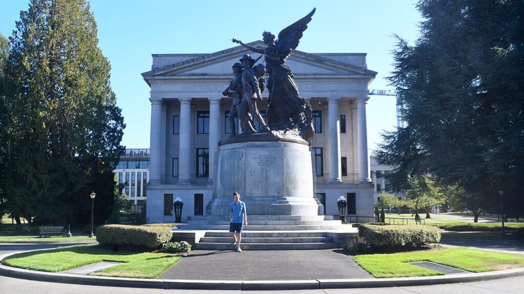 A person walks near a large statue featuring several figures in front of a neoclassical government building surrounded by trees.