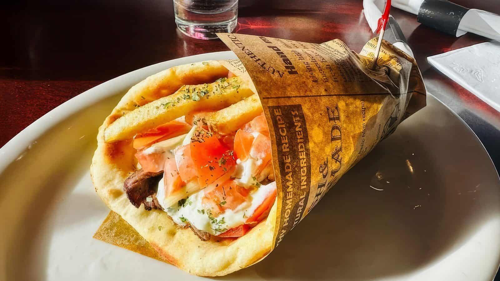 A comfort food classic, this gyro wrap is filled with sliced meat, tomatoes, onions, and tzatziki sauce, served on a white plate and partially wrapped in newspaper-style paper.