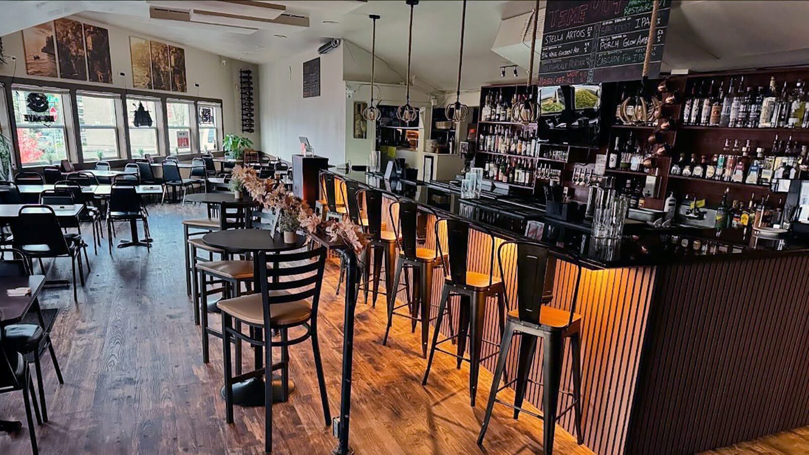 A modern restaurant interior with empty tables and chairs, a long bar with high stools, shelves of bottles, and large windows letting in natural light—perfect for enjoying comfort food like the Time Out Burger.