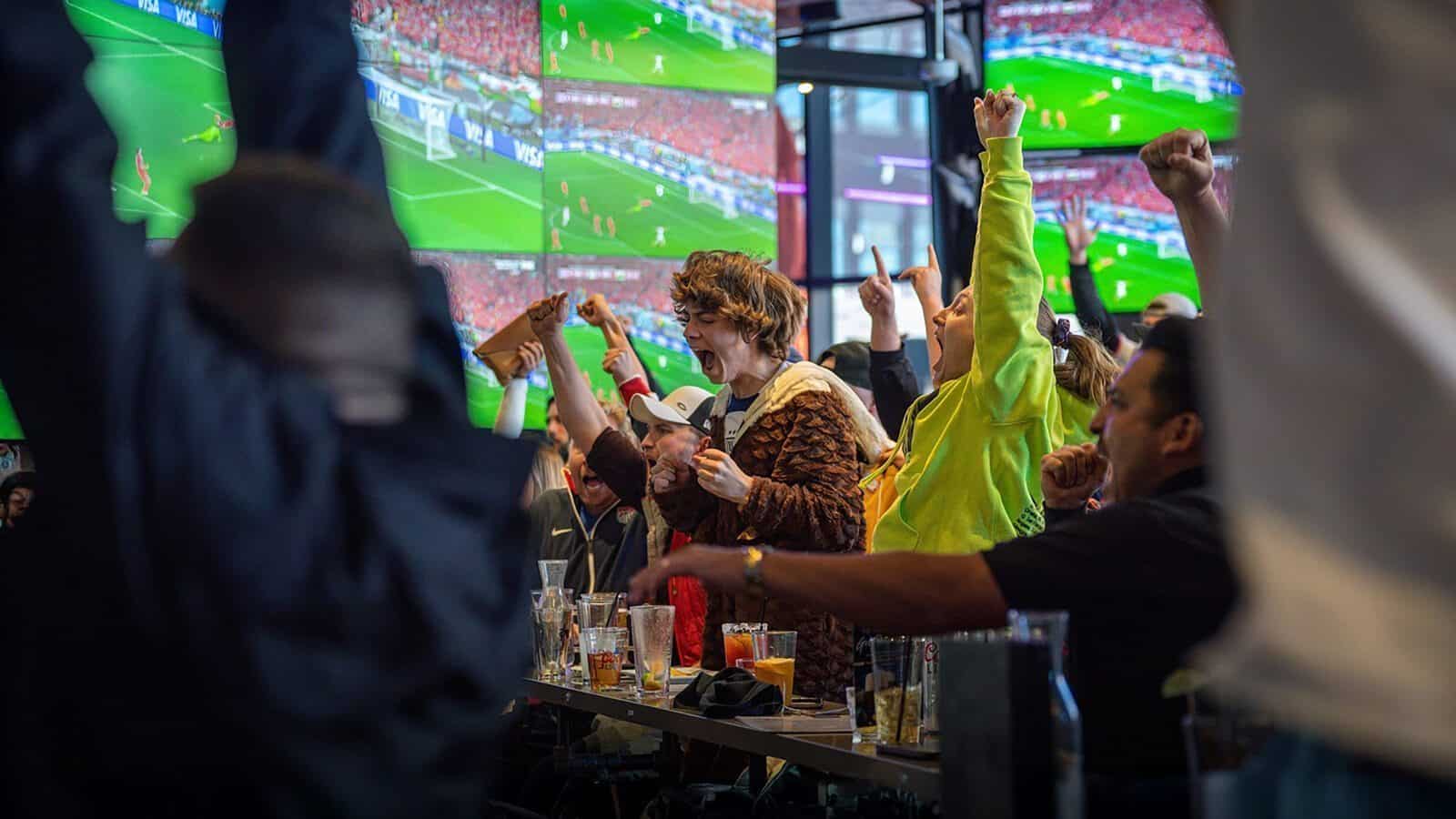 A group of people at a sports bar cheer enthusiastically while watching a soccer match on multiple large screens.