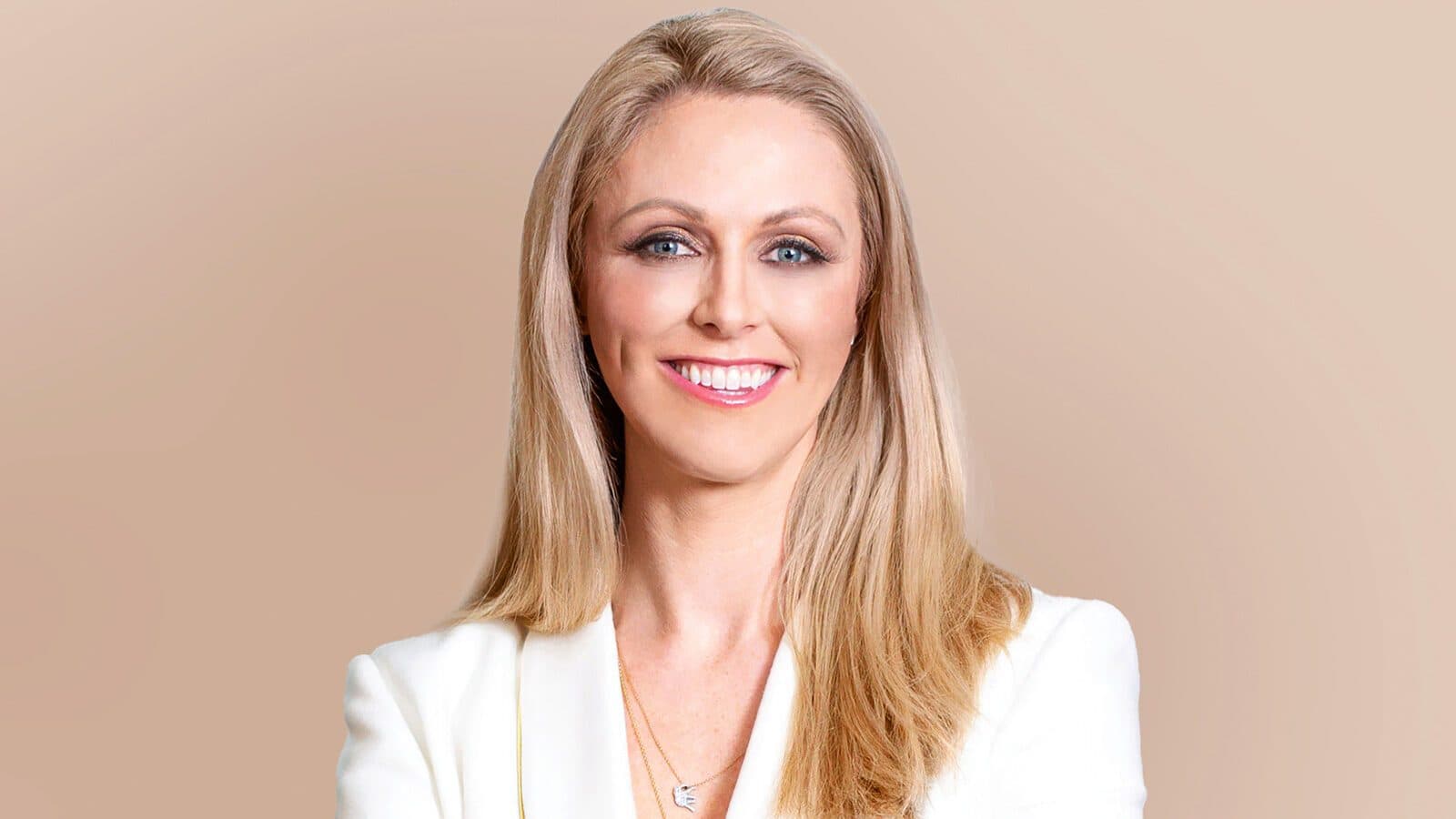 Dr. Meghan Nadeau, a woman with long blonde hair, wears a white blazer and a necklace, smiling at the camera against a plain beige background.
