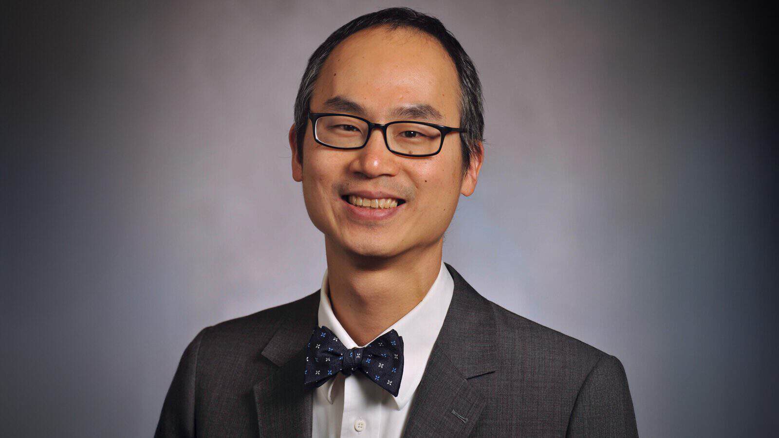 Dr. Tobias Lee, M.D., wearing glasses, a dark suit, a white shirt, and a polka dot bow tie, smiles in front of a plain background.