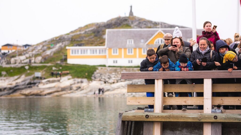 A group of people, including several children, stand on a wooden dock in Nuuk, Greenland, looking down at the water, with buildings and a hill in the background.