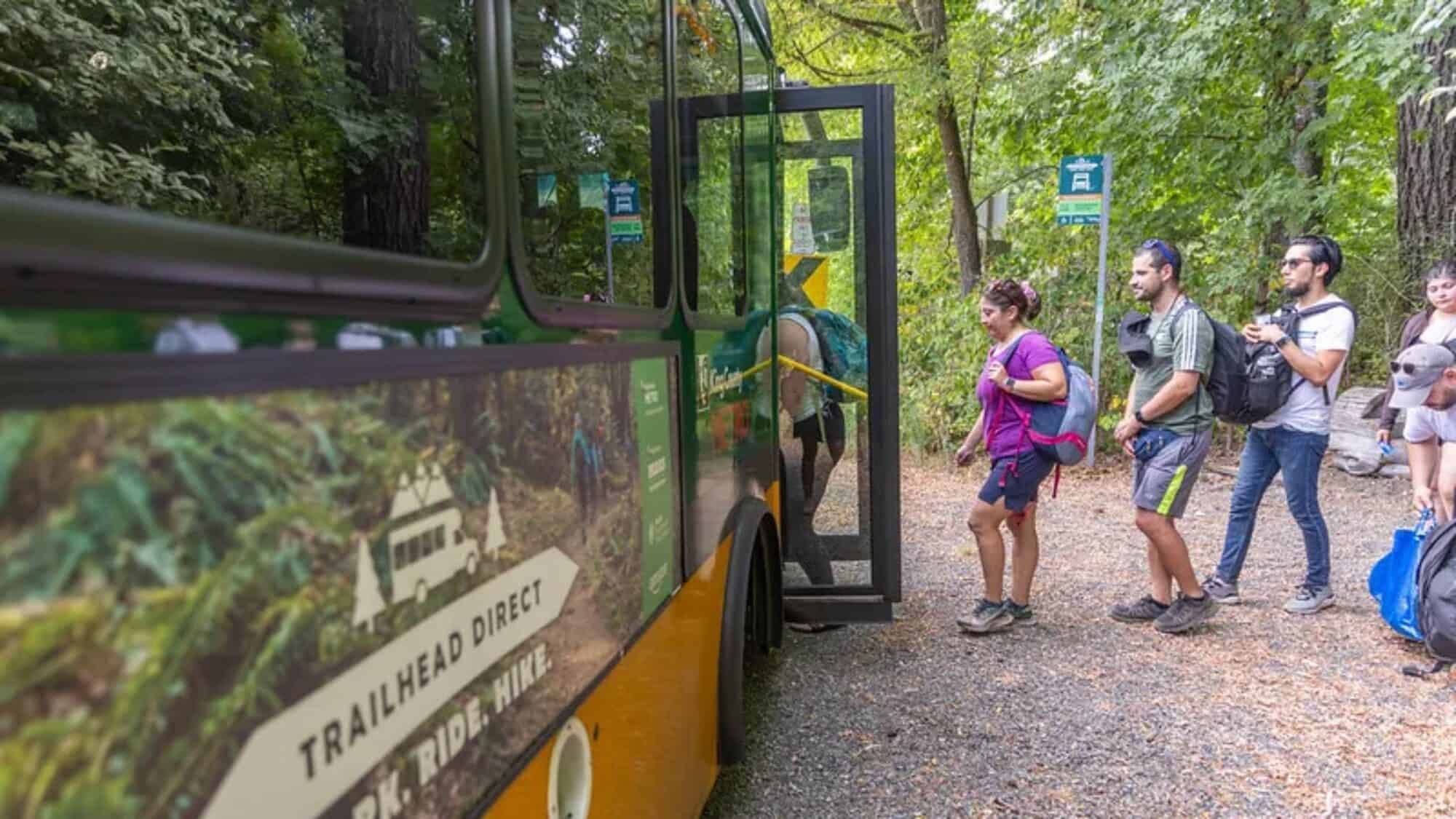 People with backpacks line up to board a green and yellow Trailhead Direct bus at a forest trailhead on a gravel path.