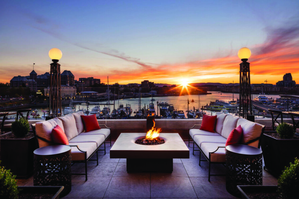 Rooftop lounge with a fire pit, cushioned seating, and a view of the marina at sunset. The sky is colorful, with the sun just above the horizon, giving way to dark skies that unveil celestial magic as stars begin to twinkle.