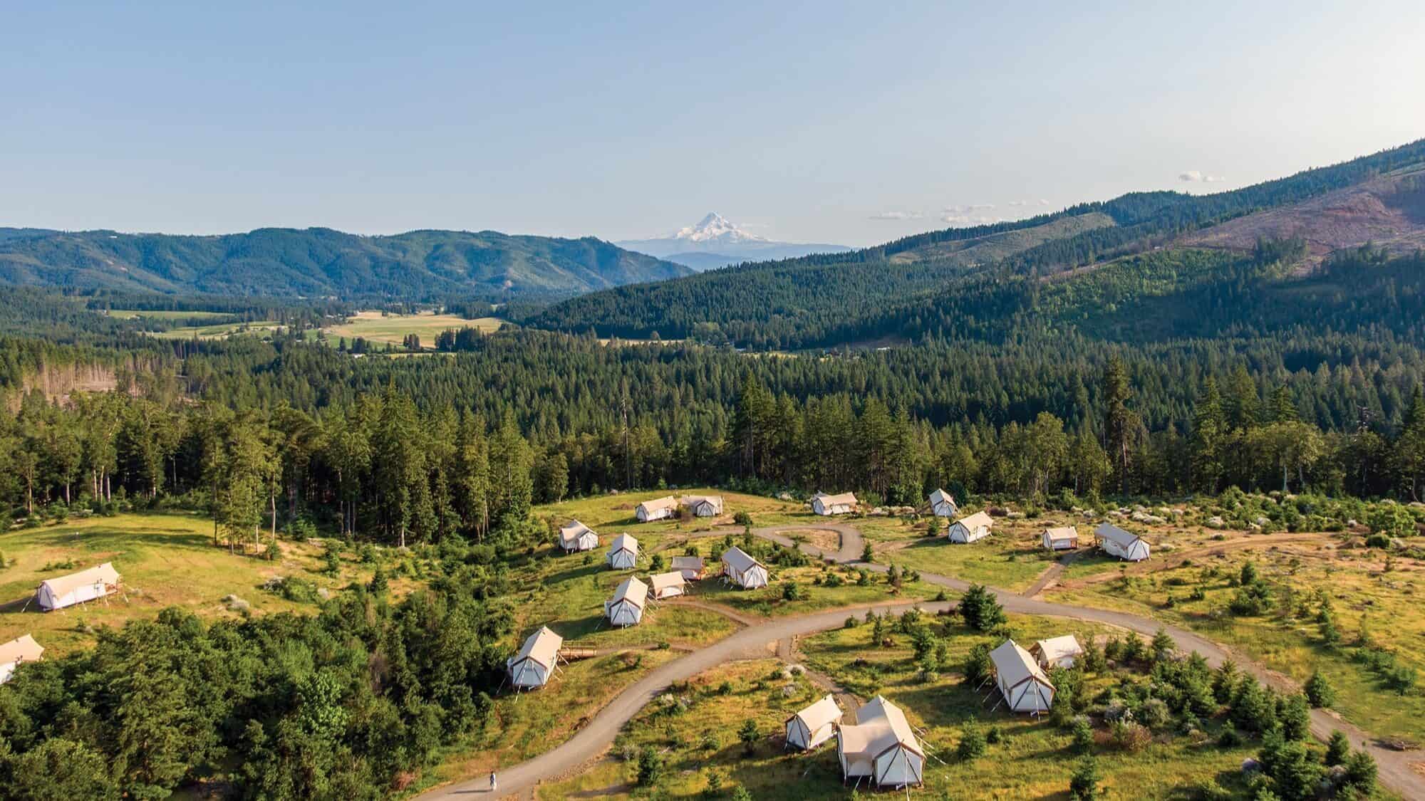 A cluster of canvas tents is set among winding paths in a grassy, wooded area, offering luxury camping with mountain views and a snow-capped peak rising in the background.