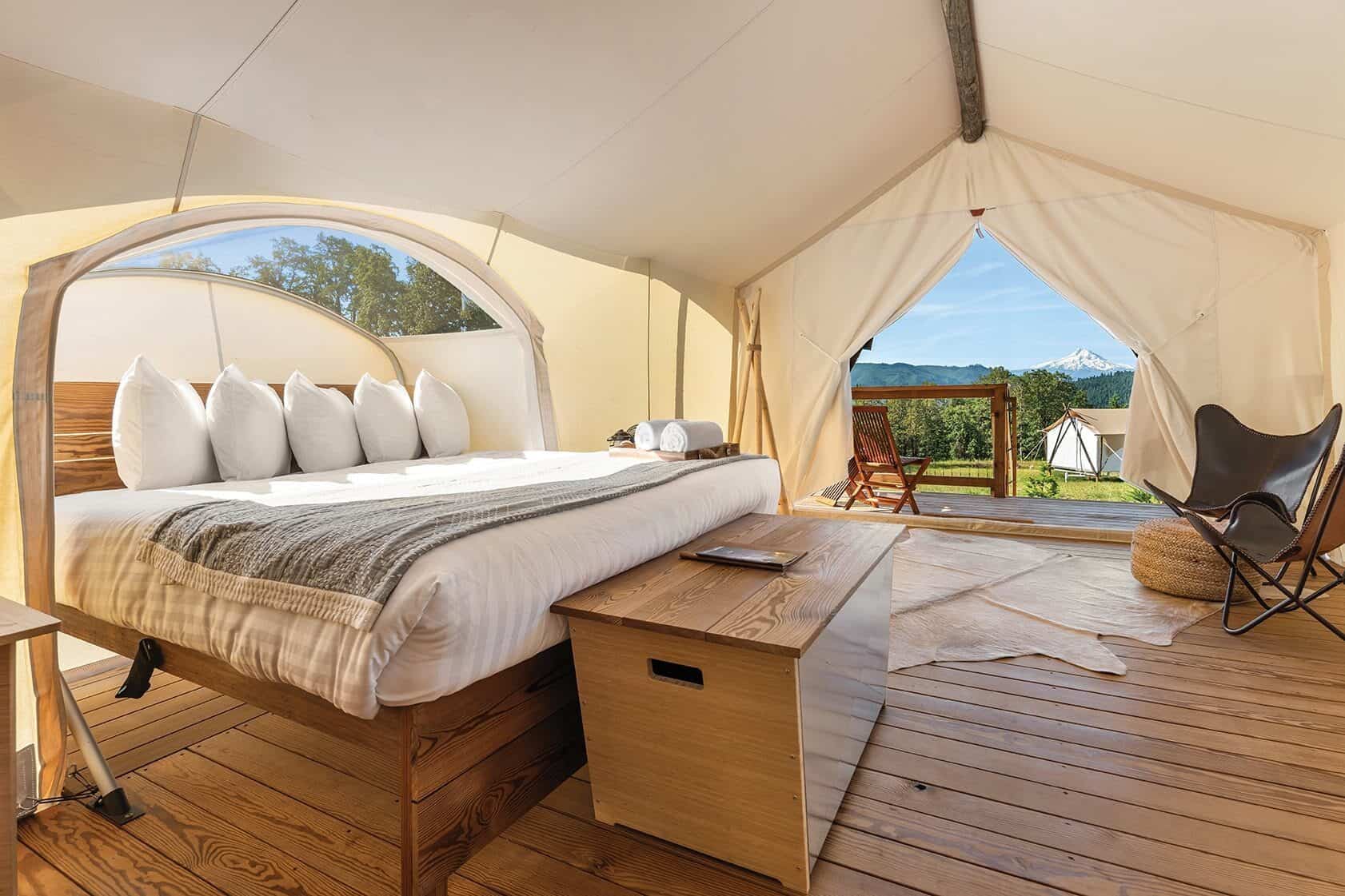 A spacious Under Canvas Northwest glamping tent features a bed with white linens and pillows, a wooden bench, a chair, and a view of greenery through the open front.
