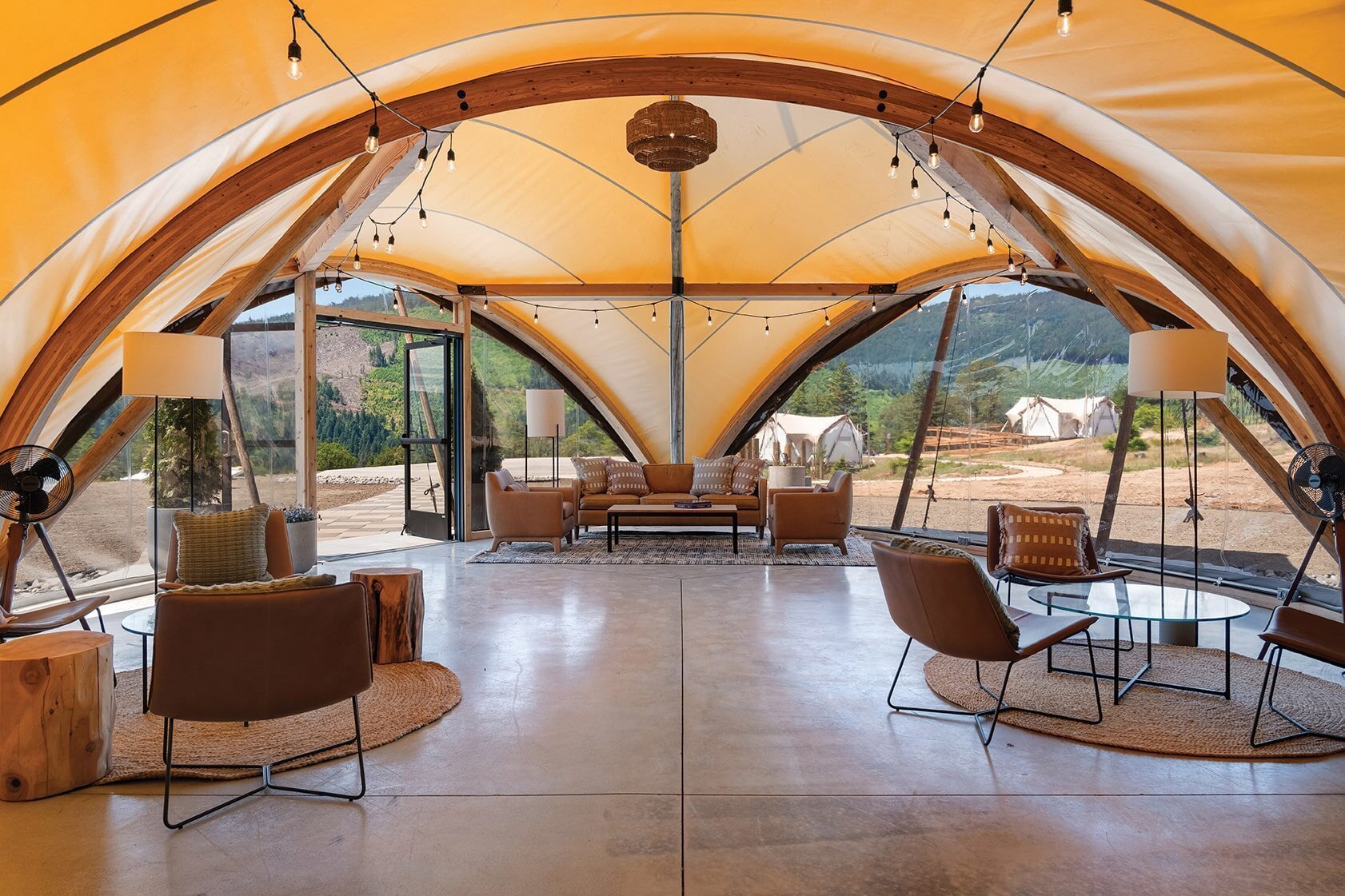 Large, modern tent with arched wooden beams, stylish seating areas, and large windows overlooking outdoor tents and a scenic mountain view—an ideal setting for glamping and upscale outdoor hospitality at Under Canvas Northwest.
