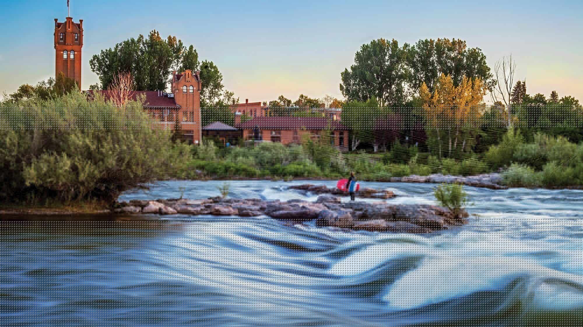 A person sits on rocks by a flowing river with historic brick buildings and trees in the background during sunset, capturing the charm of small towns and the scenic beauty that defines Montana tourism.