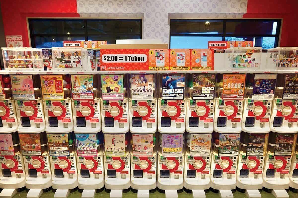A row of capsule toy vending machines with various toys on display and a sign above stating "$2.00 = 1 Token," perfect for a fun pit stop while exploring Asian gardens or looking for what to do in Seattle in the summer.