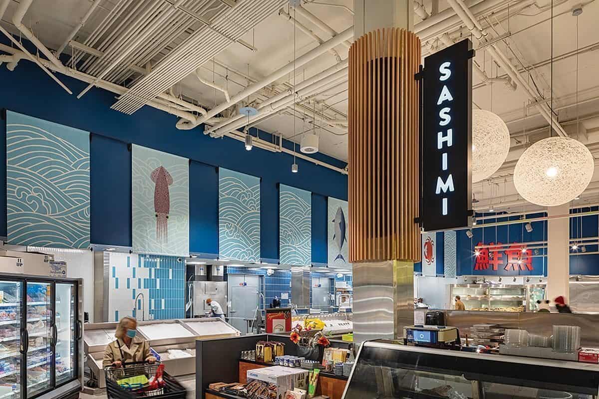 A supermarket seafood section with a prominent "Sashimi" sign, blue wave-themed wall art, and display cases—perfect inspiration for enjoying summer in Seattle, with a person shopping in the foreground.