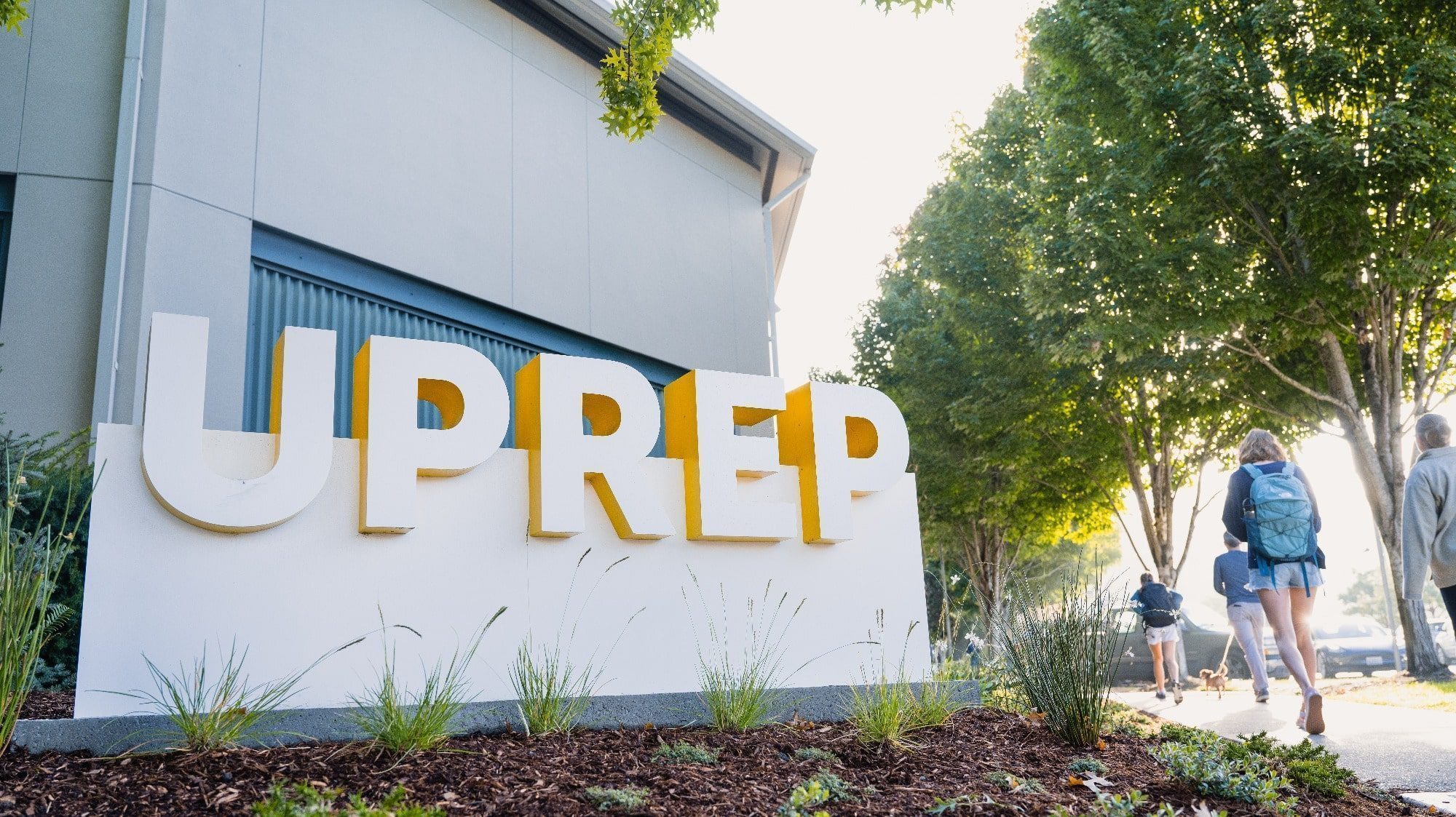Students walk near a sign that reads "UPrep" in front of a building surrounded by trees and plants, showcasing an environment that prioritizes student wellness.