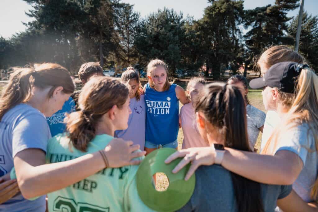 A group of people stands in a circle with their arms around each other in an outdoor setting, engaging in a discussion or pep talk. One person in the center, representing UPrep, holds a green flying disc, symbolizing their commitment to prioritizes student wellness.