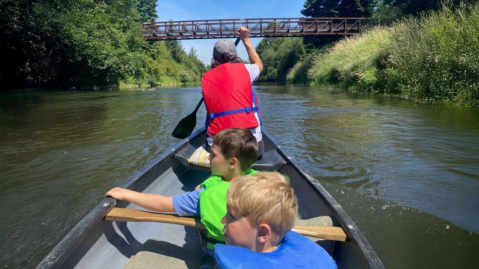 An adult and two children wearing life jackets paddle a canoe down a river toward a metal footbridge surrounded by trees and grass.
