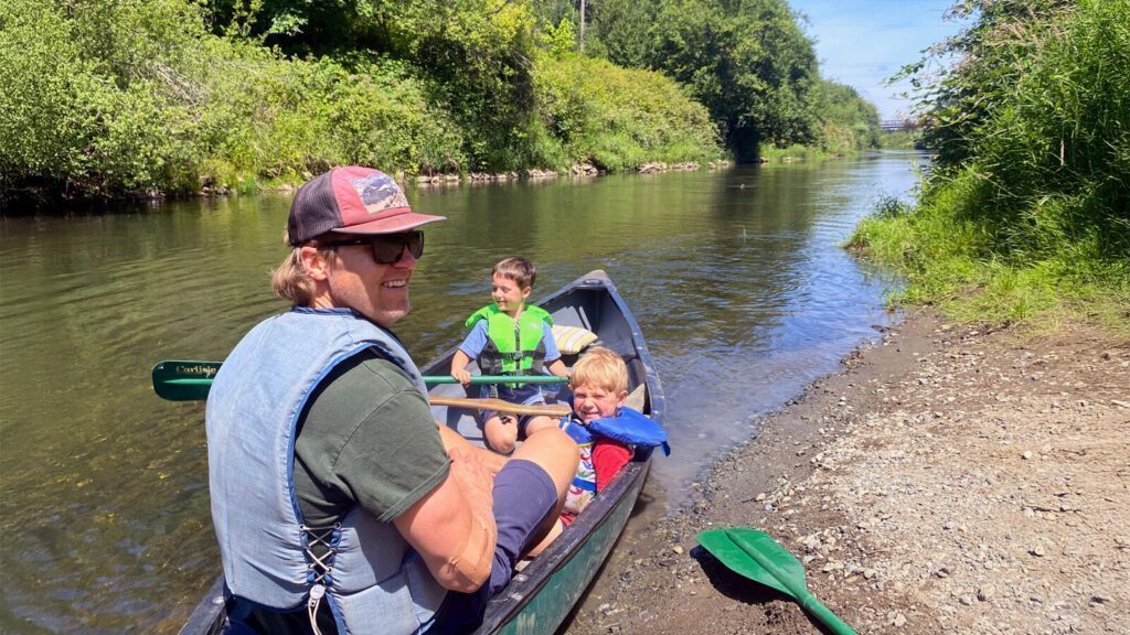 An adult and two children in life jackets sit in a canoe at the edge of a calm river, with trees and greenery along the riverbank.