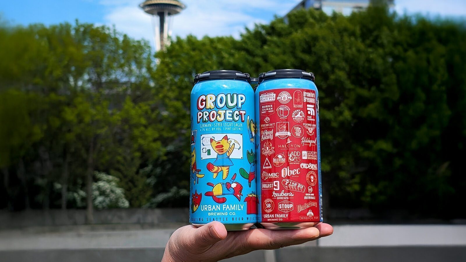 A hand holds two colorful cans of Urban Family Brewing beer outdoors, with the Space Needle and green trees in the background—perfect for celebrating Seattle Beer Week.