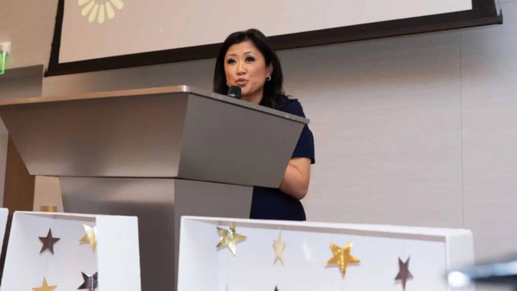 A woman stands at a podium speaking into a microphone in a modern indoor setting, with gold star decorations visible in the foreground.
