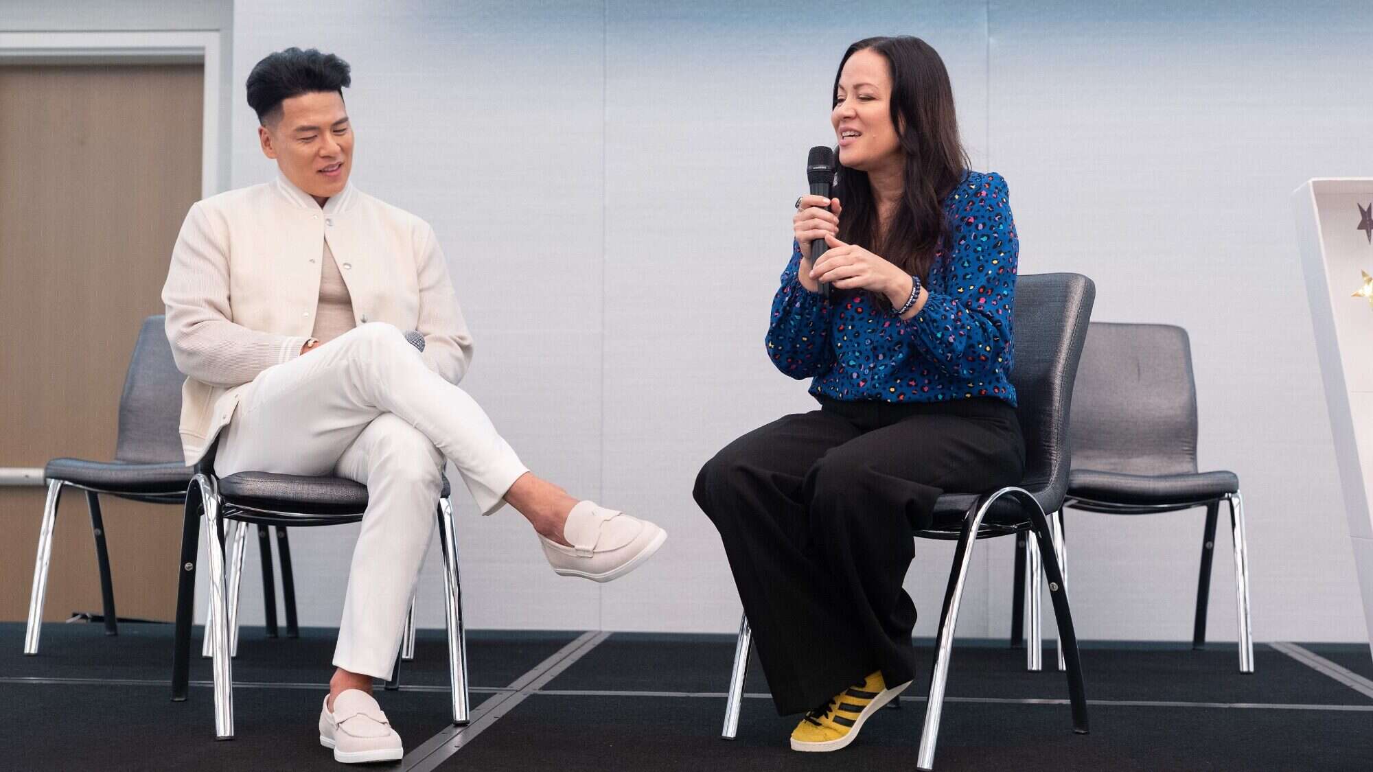 A man and a woman sit on chairs on a stage; the woman holds a microphone and speaks while the man listens attentively.