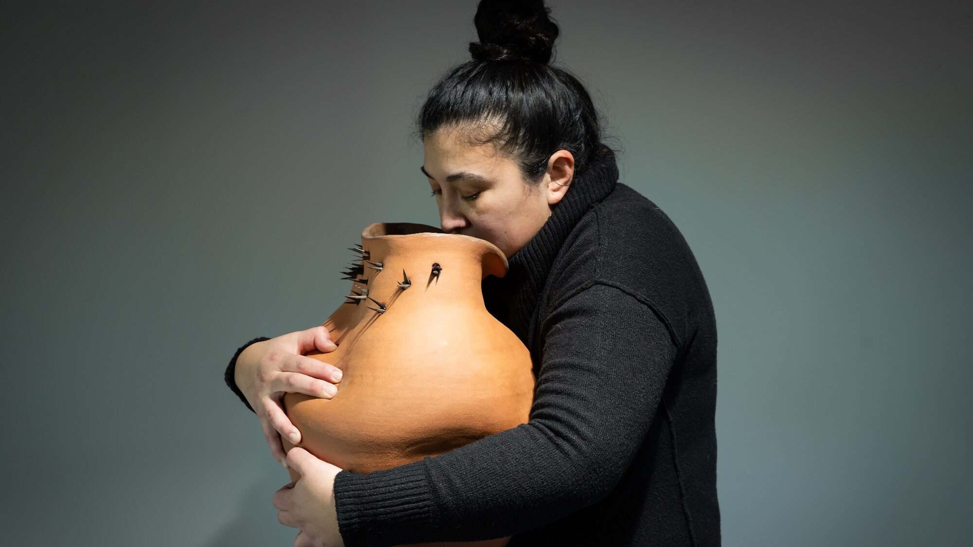Person in a black turtleneck holds and leans face against a large, irregularly shaped clay pot with cracks and small black objects attached.