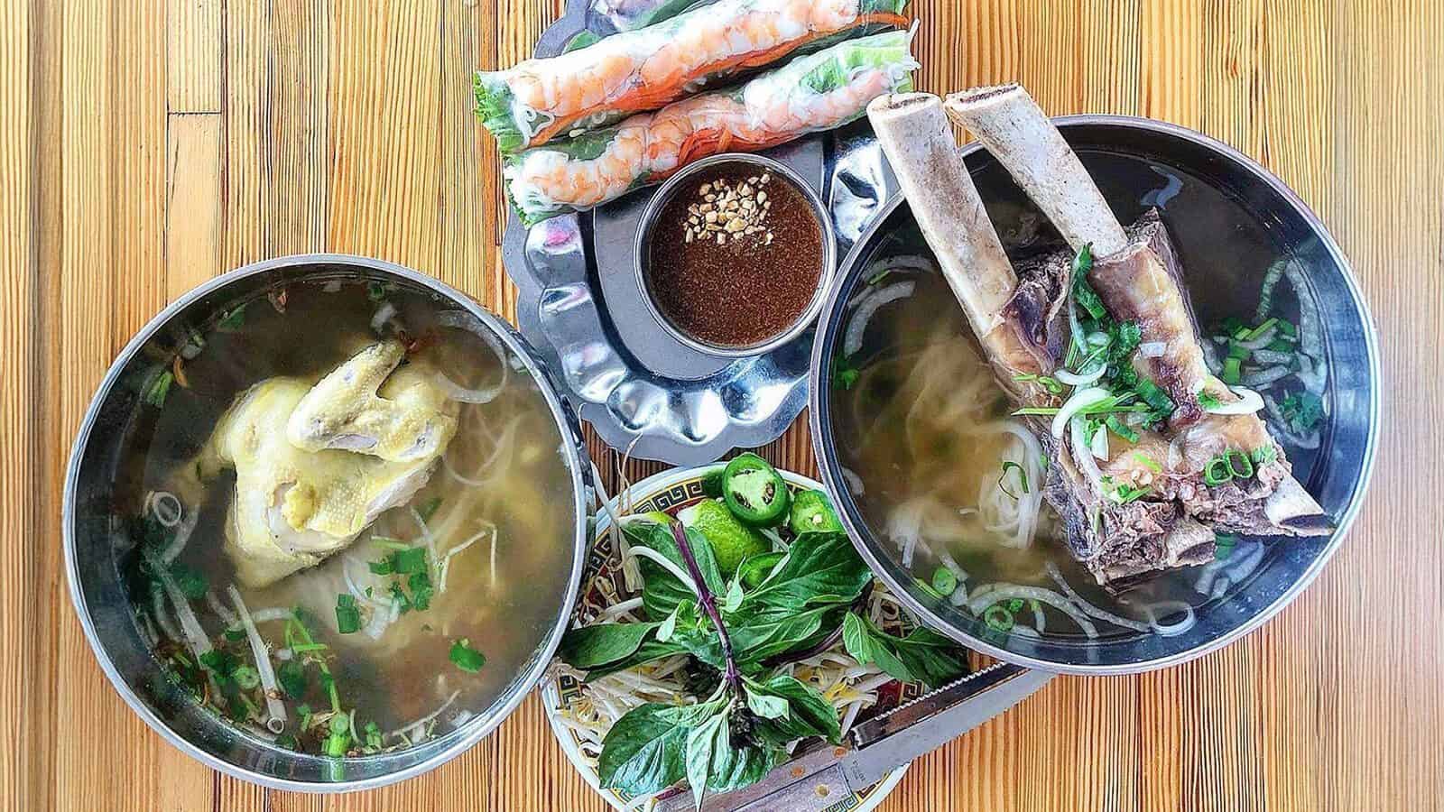 Two bowls of Vietnamese pho, one with chicken and one with beef bone, sit next to fresh spring rolls, dipping sauce, and a plate of herbs, jalapeños, and lime—a delicious nod to the AANHPI community in Seattle cultural landmarks.