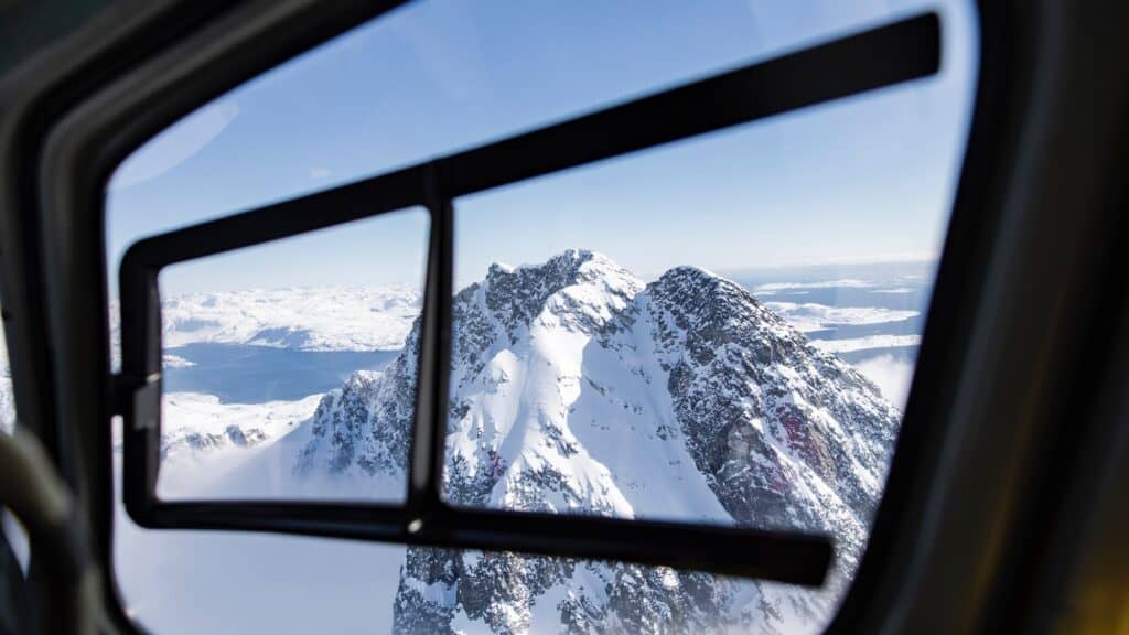 Snow-covered mountain peak viewed through an open window from inside an aircraft, with a clear blue sky and distant water in the background—an awe-inspiring glimpse of Greenland near Nuuk.