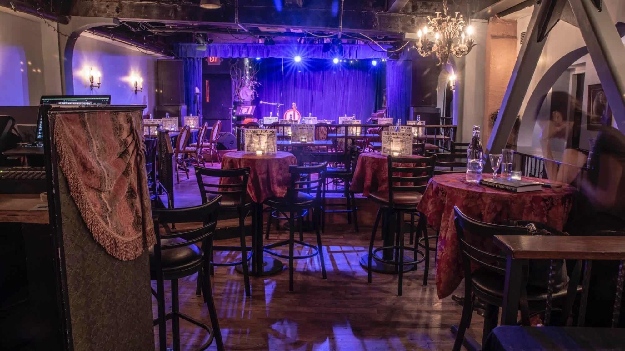 A dimly lit cabaret-style music venue near Pike Place, featuring round tables, black chairs, decorative lamps, and a stage with a blue curtain in the background.