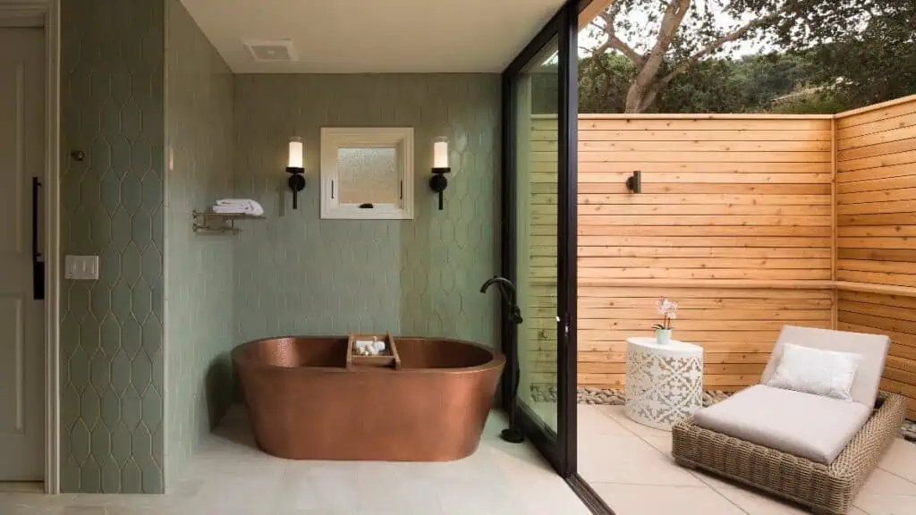 A modern bathroom with a copper bathtub, wall-mounted lights, and sliding glass doors opening to an outdoor patio with a lounge chair and small table.