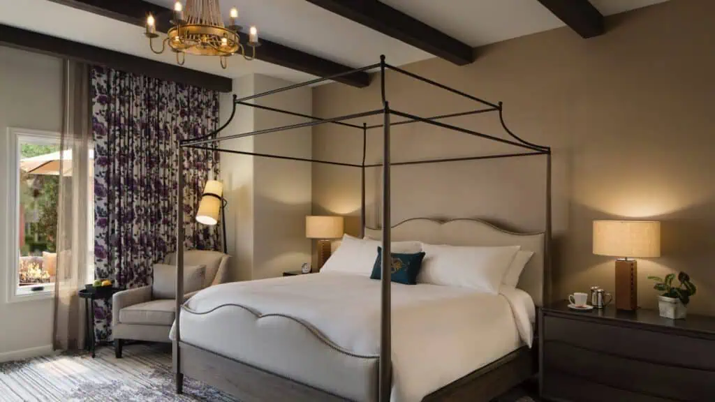A well-lit bedroom with a canopy bed, two bedside tables with lamps, an armchair by the window, and floral curtains.