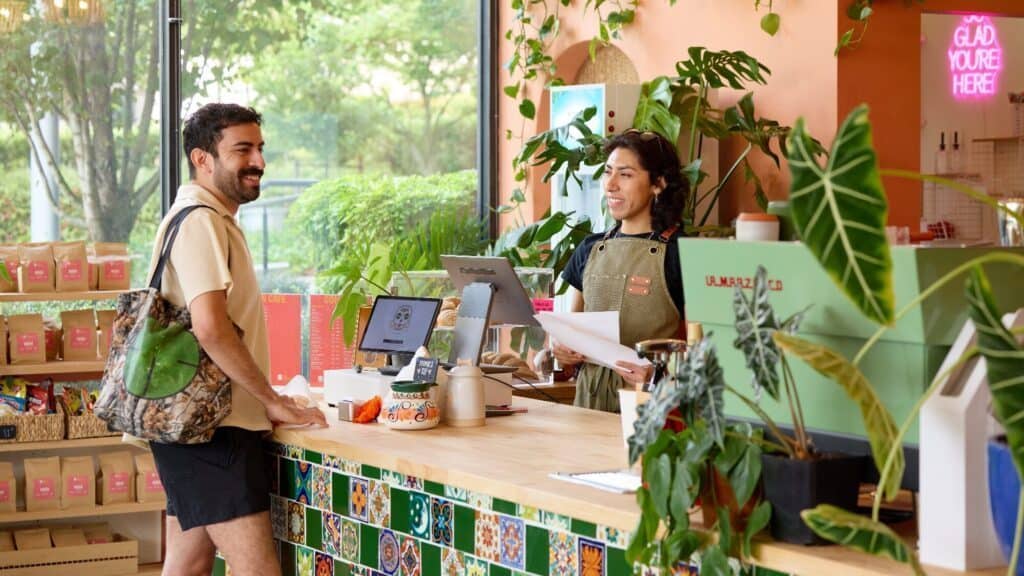 A barista stands behind a cafe counter smiling at a customer holding a bag. The counter has plants and colorful tiles, and shelves of coffee products are visible in the background.